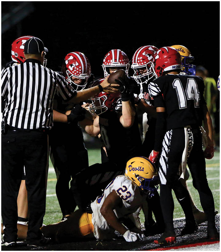 A group of football players in red and black uniforms surround a player in a white and yellow uniform who is lying on the ground near the sideline while a referee signals nearby. Description generated by AI