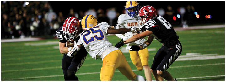 Two people in black uniforms are tackling a person in a white and yellow football uniform on a field while another person in white and yellow runs nearby. Description generated by AI