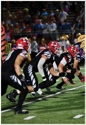 Two opposing football teams in red and black uniforms and yellow and white uniforms are lined up facing each other on a field, ready for a play. Description generated by AI