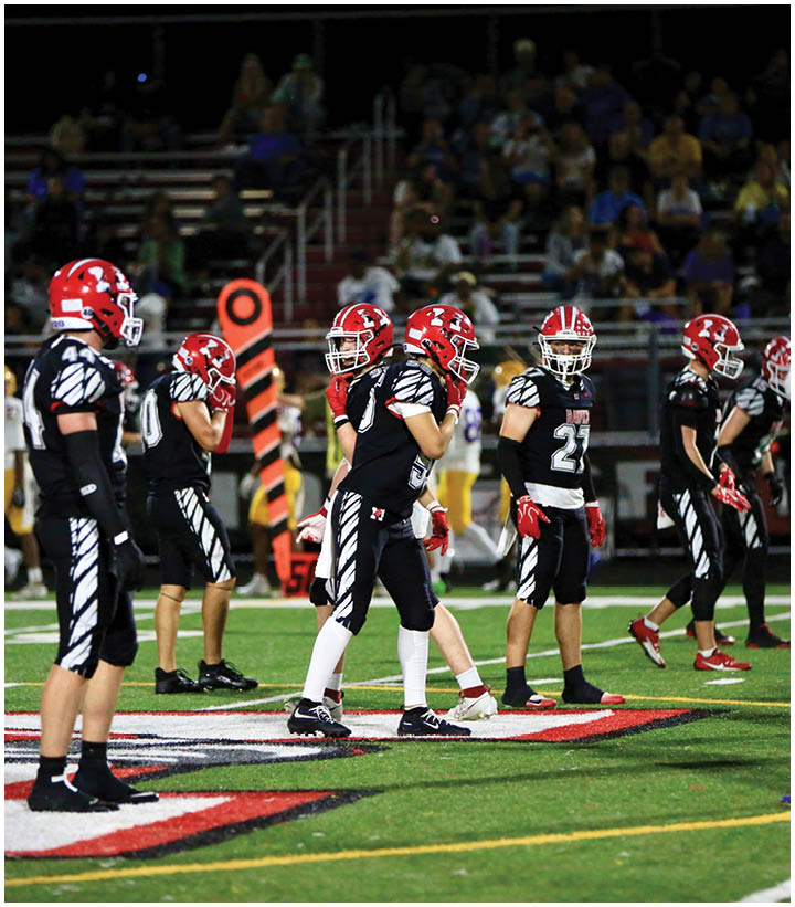 Several people wearing black and red football uniforms stand on a field facing two people in white and yellow football uniforms during a game. Description generated by AI