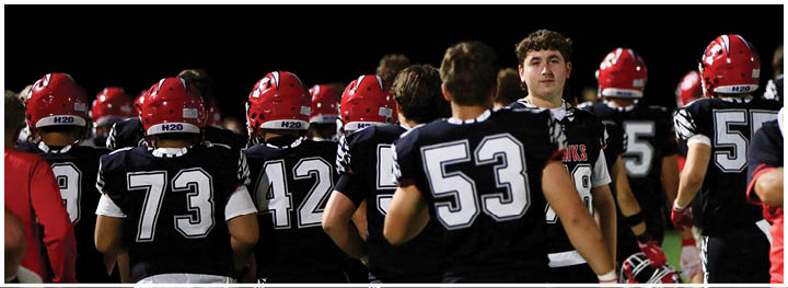 People wearing black football uniforms with red helmets are standing on a field, facing away from the camera. Description generated by AI