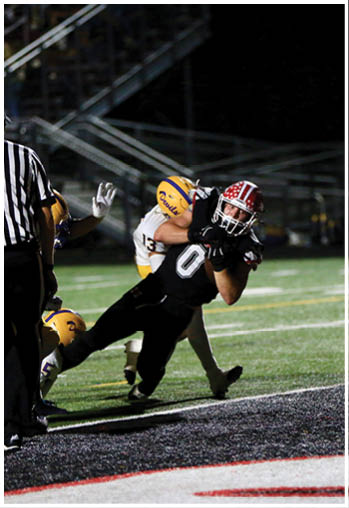 A person in a black football uniform is diving toward the end zone while being tackled by a person in a white and yellow uniform with a referee nearby on a football field at night. Description generated by AI