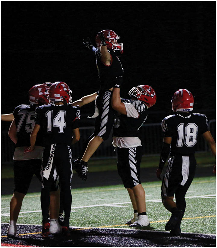 Four people in black and red football uniforms celebrate on a field at night with one person being lifted into the air by another. Description generated by AI
