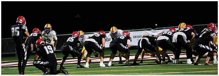 A group of football players in helmets and uniforms are lined up on a field under bright lights at night, preparing for a play. Description generated by AI