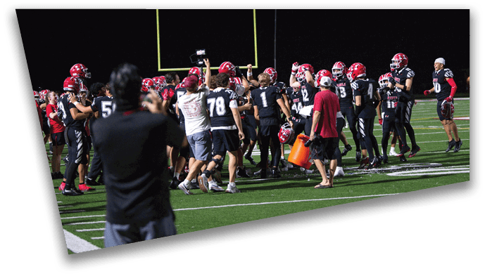 A group of people wearing football uniforms and helmets are gathered on a field at night, celebrating near the goalposts while one person in the foreground records the scene. Description generated by AI