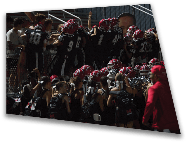 A group of people wearing football uniforms and helmets are gathered closely together near a fence while another group of people in cheerleading outfits stand nearby, with one person in a red jacket walking past. Description generated by AI