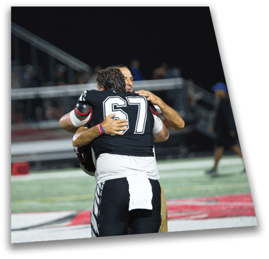 Two people in football uniforms are embracing on a field while another person in a football uniform stands nearby. Description generated by AI