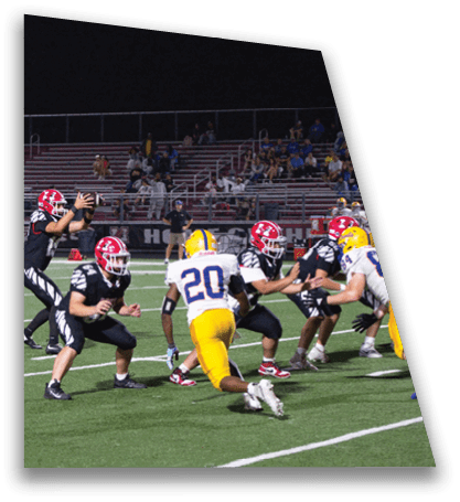 People in football uniforms are actively playing a nighttime game on a field with yard lines visible. Description generated by AI