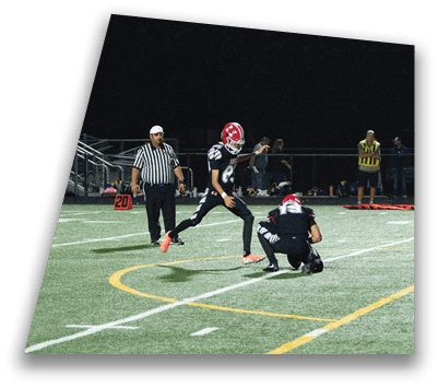 Two people in football uniforms are on a field with one holding the ball for the other to kick while a referee and spectators watch in the background. Description generated by AI