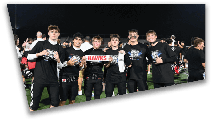 A group of six people wearing black shirts stand together on a field at night holding a sign that says “HAWKS" and smiling. Description generated by AI