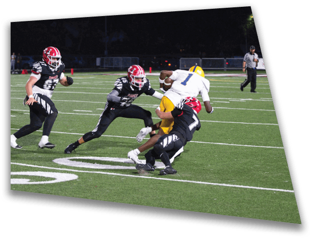 A group of people playing football on a field at night with one person in a white and yellow uniform being tackled by two people in black and red uniforms. Description generated by AI