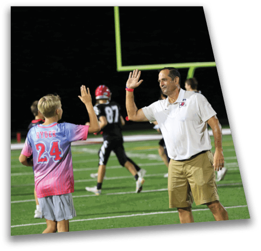 An adult wearing a white shirt and khaki shorts is giving a high five to a child in a pink sports jersey with the name Ryder and number 24 on a football field at night. Description generated by AI