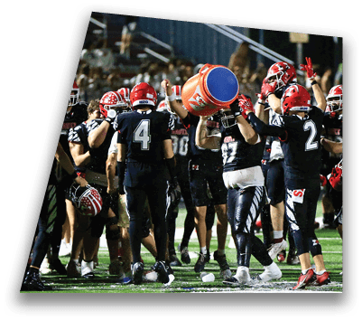 A group of football players in black uniforms with red helmets celebrate on a field as one player pours water from a large orange cooler over another. Description generated by AI