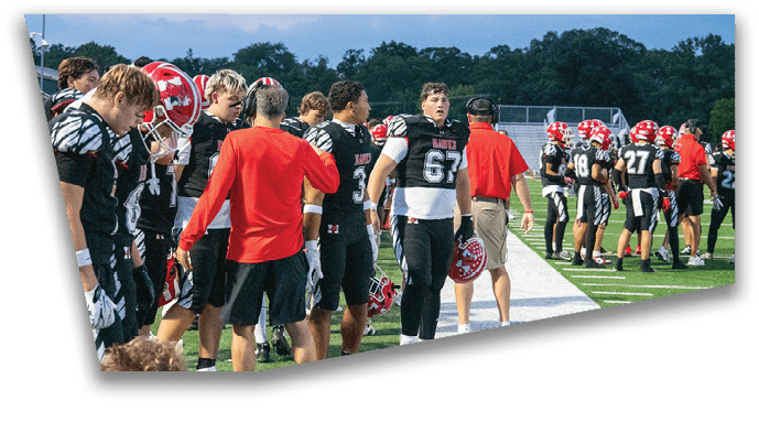 A group of people wearing football uniforms and helmets stand on a field with some facing forward and others turned away under a cloudy sky. Description generated by AI