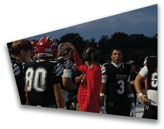 People in football uniforms and a person in a red shirt are gathered outdoors during dusk, appearing to prepare or discuss something. Description generated by AI