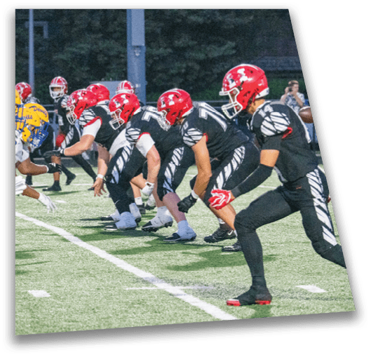 A group of people wearing black sports uniforms with red helmets are lined up in a crouched position on a football field, preparing for a play. Description generated by AI
