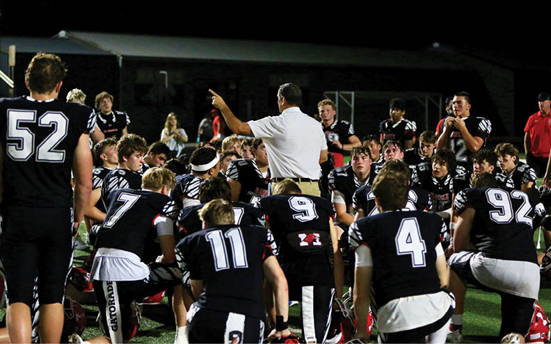 A group of people in black sports uniforms with white numbers are gathered on a field at night, listening to a person in a white shirt who appears to be giving instructions. Description generated by AI