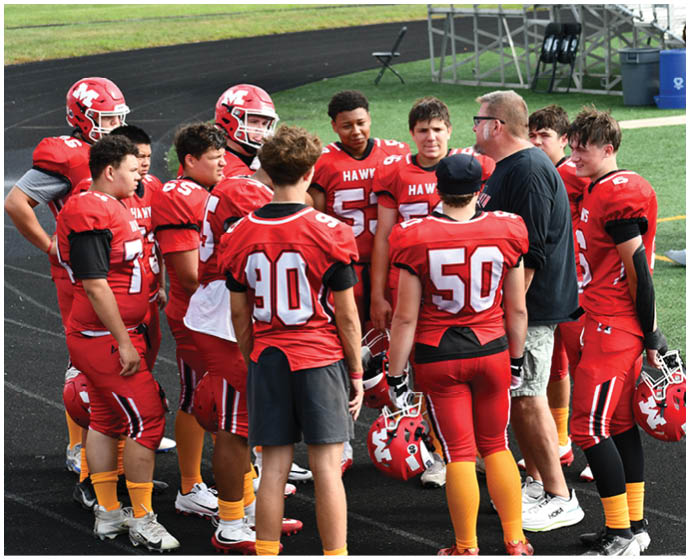 A group of people wearing red football uniforms and yellow socks are standing together on a black surface, with one person holding a red helmet. Description generated by AI