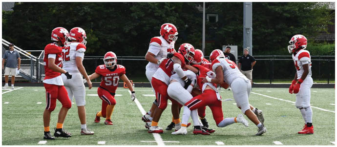 A group of people wearing red and white football uniforms are engaged in a play on a grassy field with a building and trees in the background. Description generated by AI