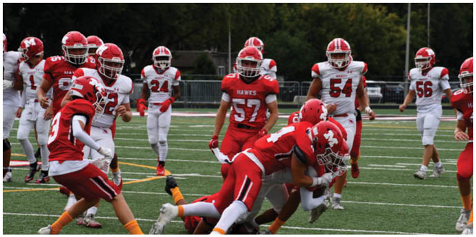 Players in red and white football uniforms are actively engaged in a tackle on a green field during a game. Description generated by AI