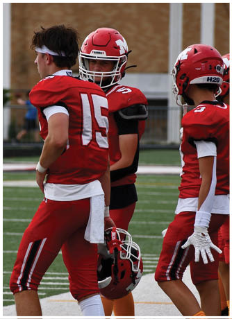 Two people wearing red football uniforms are standing close together on a field, with one wearing a helmet and the other wearing a headband. Description generated by AI
