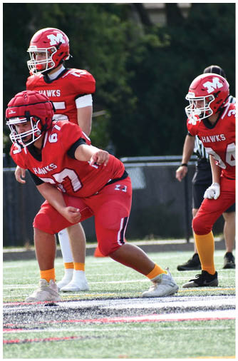 A person in a red football uniform with the number 34 is in a ready stance on a football field with another player partially visible and a referee in the background. Description generated by AI