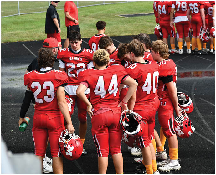 A group of people wearing red football uniforms with helmets in hand are gathered on a track near a grassy field. Description generated by AI