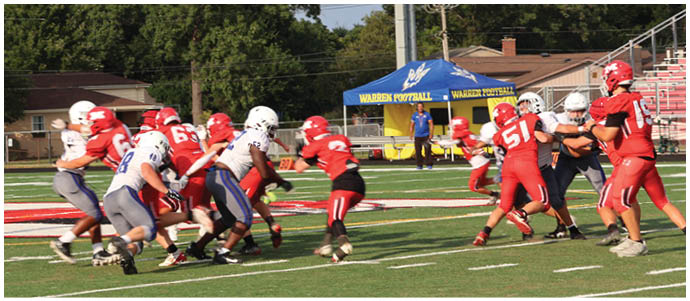 A group of people wearing football uniforms are engaged in a play on a grassy field with a referee and a blue tent visible in the background. Description generated by AI