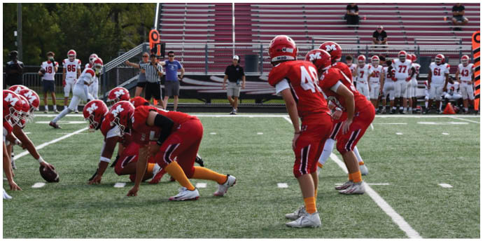 Players in red and white football uniforms are lined up on a green field preparing for a play with spectators and team members in the background. Description generated by AI
