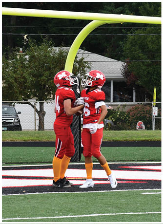 Two people in red football uniforms and helmets stand close together on a football field near the goalpost. Description generated by AI