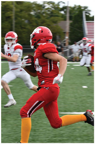 A person in red sports gear is running with a football while another person in white sports gear is chasing them on a football field. Description generated by AI