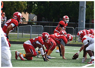 Two opposing American football teams in red and white uniforms are lined up at the line of scrimmage on a green field with a player in red positioned behind the center ready to receive the ball. Description generated by AI