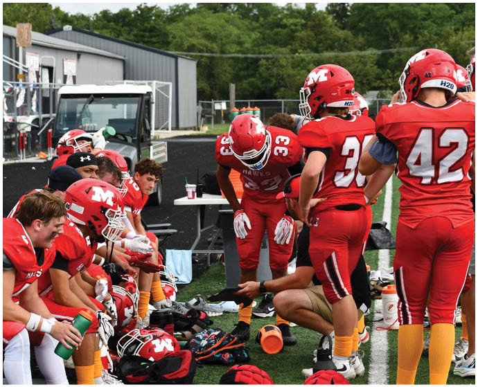 A group of people in red football uniforms are gathered on the sidelines of a field, with one person bending forward and another kneeling while holding a tablet. Description generated by AI