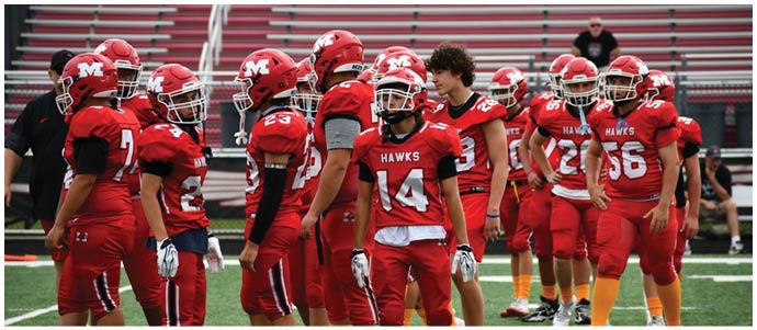 People wearing red sports uniforms and orange socks are standing on a green turf field, likely preparing for a game or practice. Description generated by AI