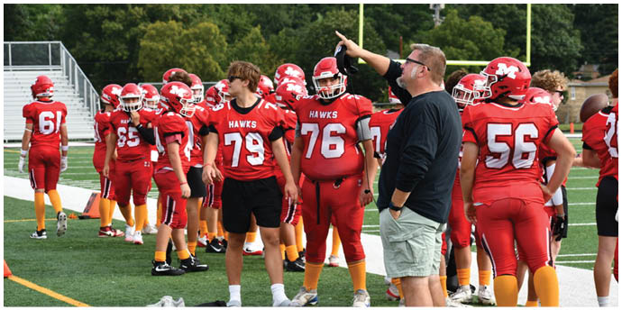 A group of people wearing red football uniforms with “HAWKS" on the jerseys are gathered on a field while one person in casual clothing appears to be giving instructions. Description generated by AI