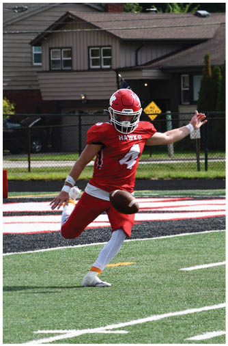 A person wearing a red football helmet and jersey with the word “HAWKS" is standing in front of a house with beige siding and a brick chimney. Description generated by AI