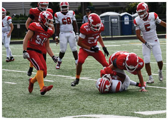 Four people in football uniforms are on a field with one person on the ground being tackled by another while two others stand nearby. Description generated by AI