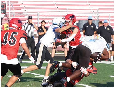 Players in red and white football uniforms are engaged in a tackle on a green field with empty bleachers and several people standing in the background. Description generated by AI