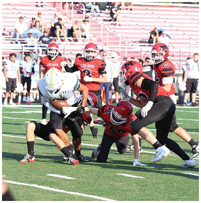 A group of people in football uniforms are actively engaged in a game on a field with bleachers and spectators in the background. Description generated by AI