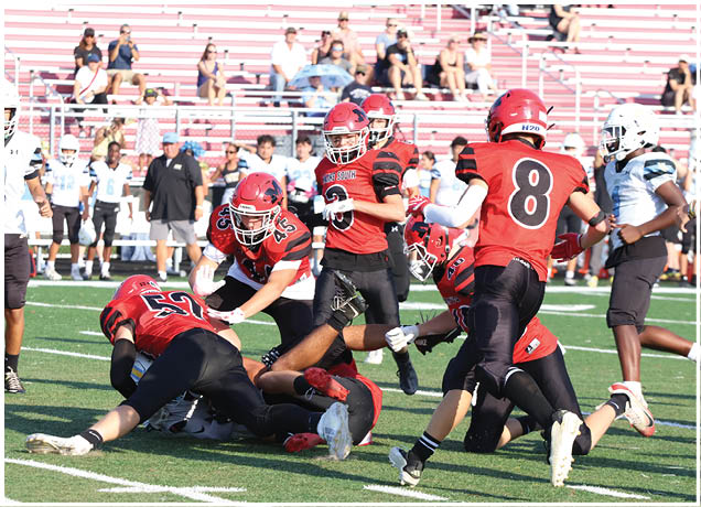 Several people wearing red and black football uniforms are engaged in a play on a green field with spectators in the background. Description generated by AI