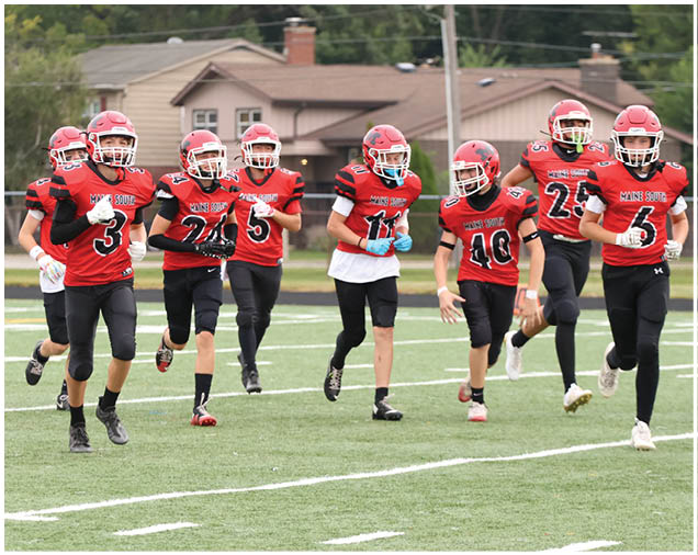A group of people wearing red football uniforms and helmets are running together on a green football field with houses and trees in the background. Description generated by AI