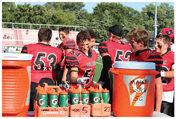 Several people wearing red and black football uniforms with face paint are gathered on a sports field near orange water coolers. Description generated by AI