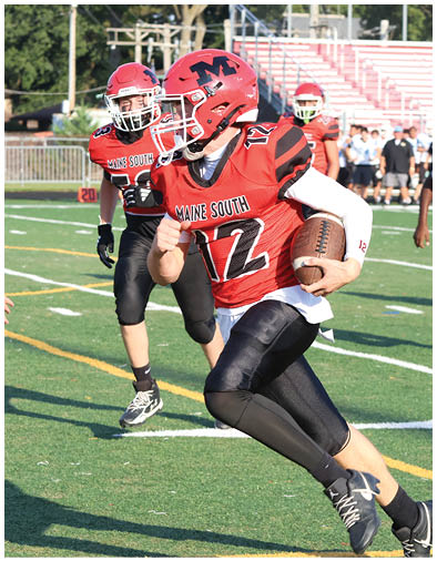 Two people in red and black football uniforms with helmets are playing on a green field, with one holding a football and running while the other follows behind. Description generated by AI