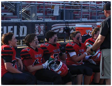 Several people wearing red and black football uniforms with face paint are sitting on a bench, holding helmets and water bottles, while interacting with someone standing nearby. Description generated by AI