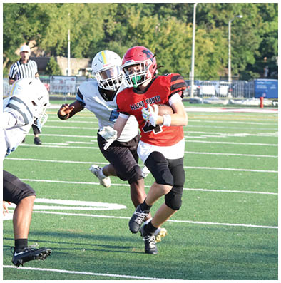 A person in a red football uniform is running with the ball while two people in white uniforms attempt to tackle them on a football field. Description generated by AI