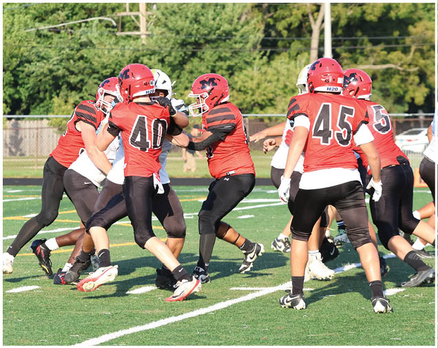 Several people wearing red and black football uniforms are engaged in a play on a grassy field with a fence and trees in the background. Description generated by AI