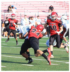 Players in red and white football uniforms are engaged in a tackle on a green field with empty bleachers in the background. Description generated by AI