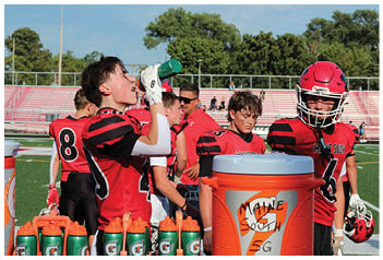 People wearing red and black sports uniforms are gathered on a sideline, with one person drinking from a green water bottle and others standing nearby, some looking focused or tired. Description generated by AI