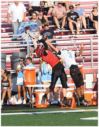 Two people stand on a sports field near a chain-link fence and bleachers, with one wearing a black shirt and gray pants and the other wearing a yellow and black striped vest, while three people sit on the bleachers in the background. Description generated by AI