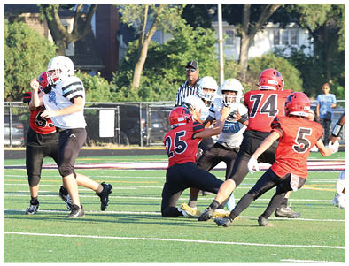Several people wearing football uniforms are actively engaged in a play on a grassy field with a referee observing in the background. Description generated by AI